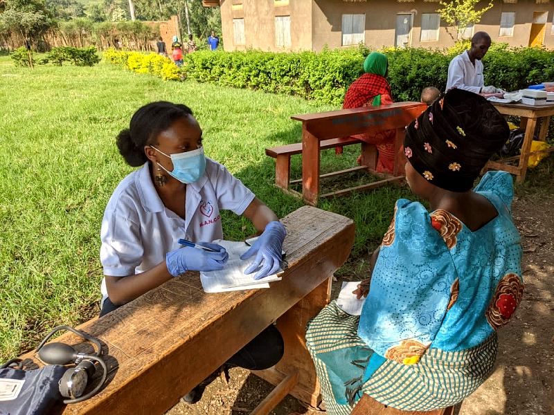 A masked MANCO midwife writes notes at an outdoor desk