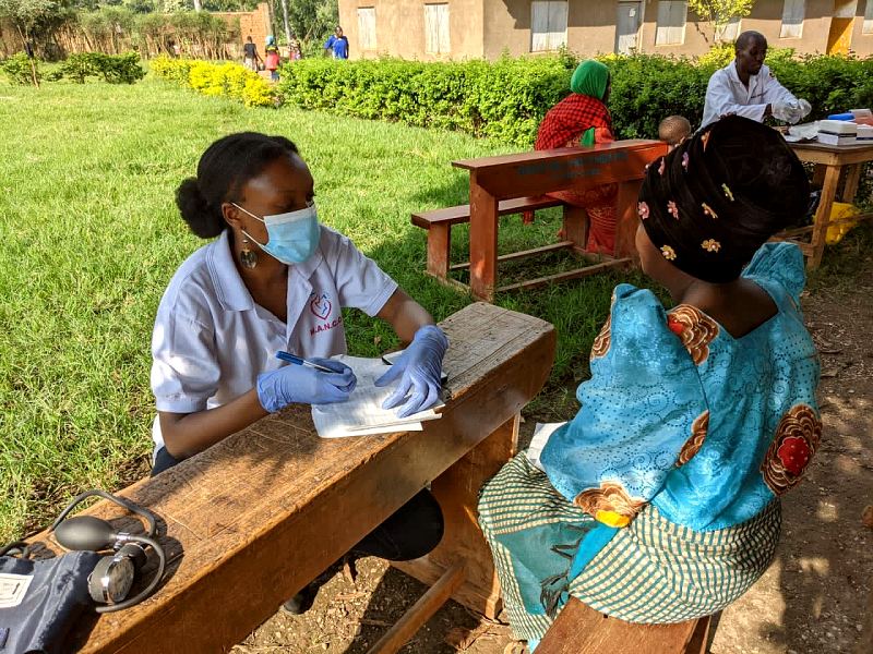 A MANCO midwife in gloves and a face mask writes notes as she talks with a mother at an outdoor clinic desk