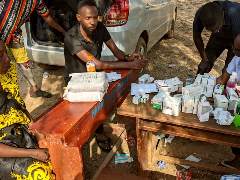 MANCO nurses run a pharmacy desk from the open boot of a car while a woman in a yellow dress waits on a bench